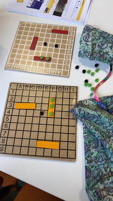 Deux plateaux de jeu quadrillés en bois posés sur une table blanche, avec des cases repérées par lettres et chiffres. Des pièces rectangulaires colorées et de pions ronds noirs et verts sont disposés sur les grilles. A côté, un pochon en tissu à motifs contient d'autres pions éparpillés.
