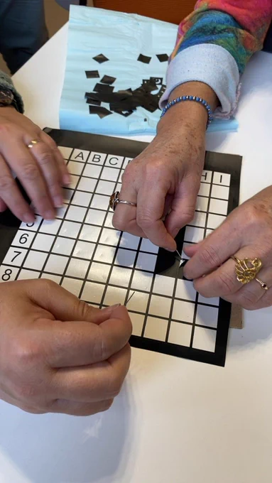 Plusieurs mains échenillent une pièce noire sur un plateau quadrillé marqué par des lettres (A à I) et des chiffres (1 à 9), en retirant délicatement les petites parties autour de la forme découpée. D'autres chutes de vinyle noir sont visibles sur la table à côté. 