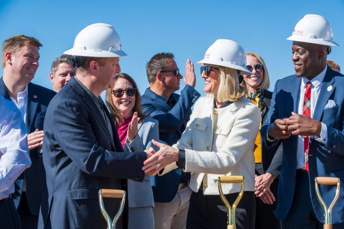 Group of professionals wearing hard hats shaking hands at a groundbreaking ceremony on a clear day.