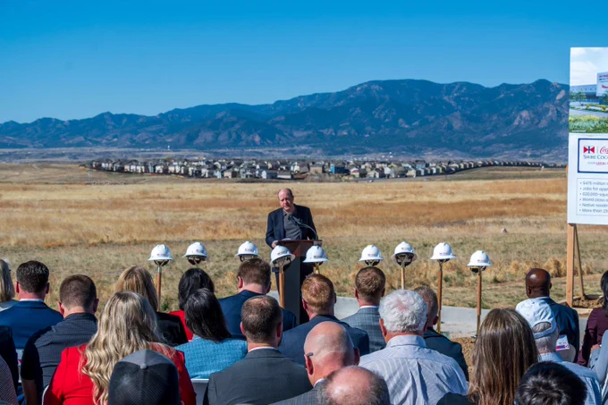 Governor Jared Polis is addressing a seated audience at a groundbreaking ceremony with mountains and suburban homes in the background.