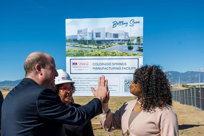 Governor Polus is giving a high-five in front of a sign announcing a new Coca-Cola manufacturing facility in Colorado Springs.