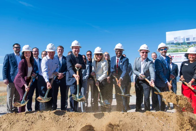 Formal groundbreaking event with people in suits and hard hats digging into soil for a new project launch, including Governor Polis and Mayor Yemi.