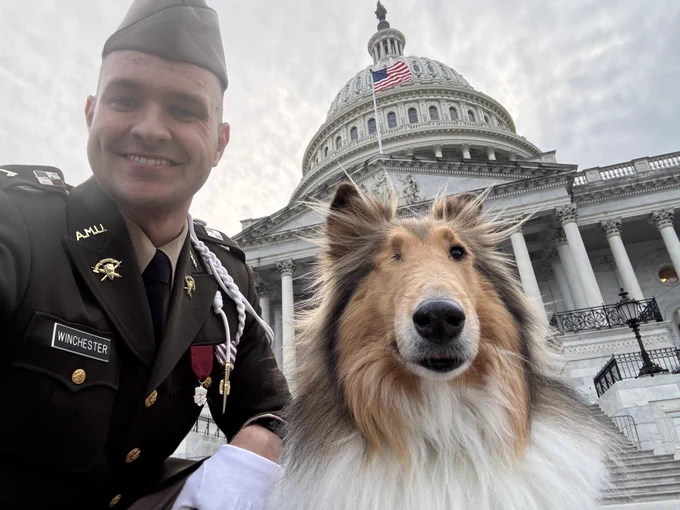Reveille's handler in a military uniform poses with Reveille the dog outside the United States Capitol building. The Capitol's dome and an American flag are visible in the background.