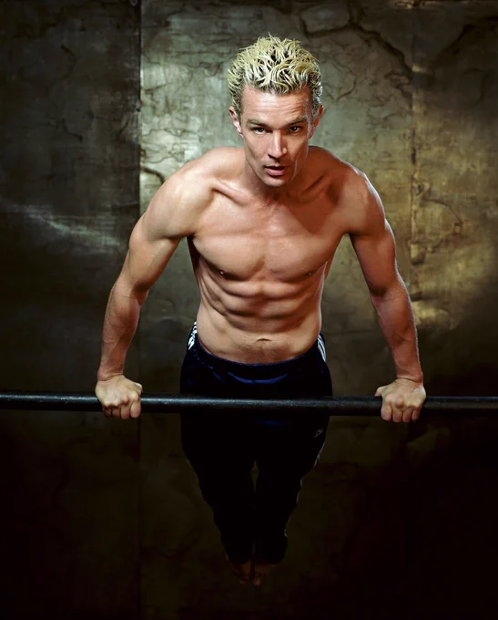 The image depicts a shirtless man performing a horizontal bar exercise. He is positioned at the center of the frame, holding onto a metallic bar with both hands. The man's arms are flexed, supporting his elevated body parallel to the bar. He has short, spiky blonde hair and a muscular build. He is wearing dark athletic pants. The background is a textured wall with a muted brown tone, adding depth to the image. The lighting highlights the contours of his muscles, emphasizing his physical form.
