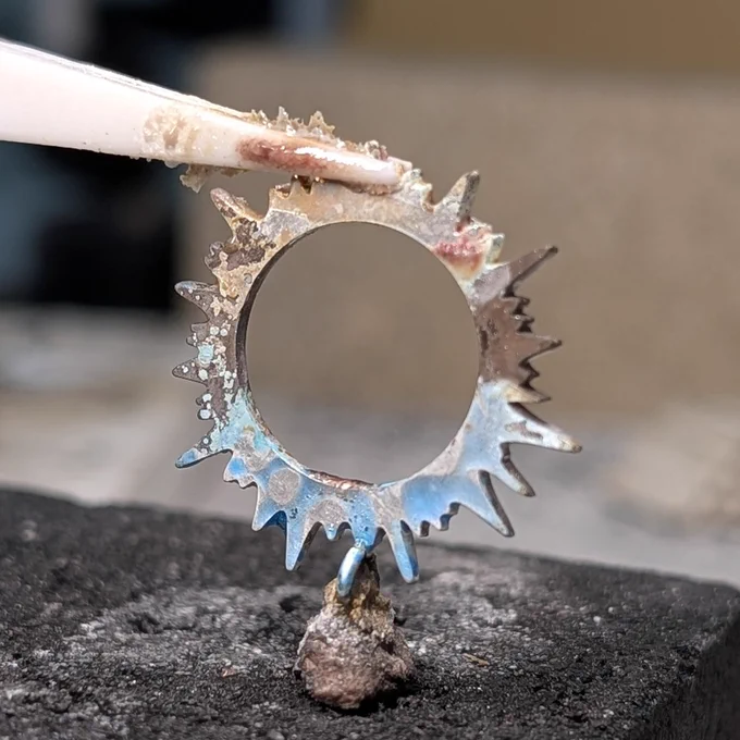 Sterling silver eclipse corona pendant mid-solder on a charcoal block, showing flux residue and oxidation on the piece.