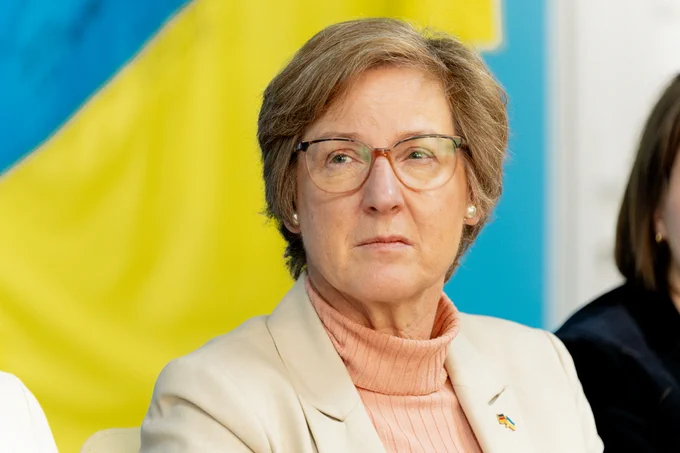 Julia Monar sits in a meeting room, listening attentively during a visit to a veterans’ centre in Ivanivka, Chernihiv Oblast. She wears glasses and a light-coloured blazer with a small pin. Behind her, a large Ukrainian flag in blue and yellow fills the background.