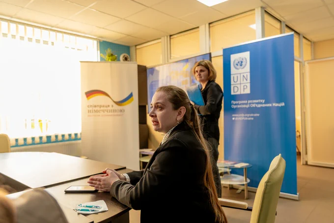 Ivanivka Village Head Olena Shvydka speaks while seated at a table during a meeting at a veterans’ centre supported by Germany and UNDP in Ivanivka, Chernihiv Oblast. Behind her are UNDP and Germany partnership banners, and another woman stands nearby holding a folder.