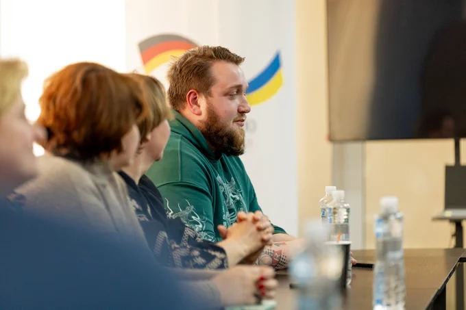 A group discussion at a meeting table inside a veterans’ centre in Ivanivka, Chernihiv Oblast. A man in a green sweatshirt speaks while others sit beside him, listening closely. 