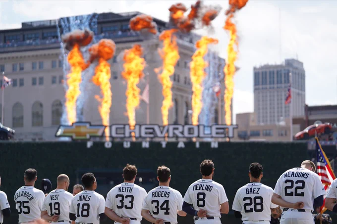 Tigers players line up on the foul line during the national anthem as flames shoot out beyond the outfield wall. They wear home white Tigers uniforms with navy blue lettering.