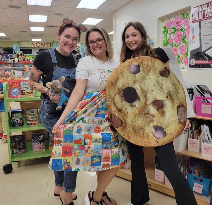 Three female educators smiling in a school library while dressed as storybook characters. One wears mouse ears and denim overalls, the center educator wears a skirt featuring classic children's book illustrations, and the third holds a large, circular chocolate chip cookie prop.