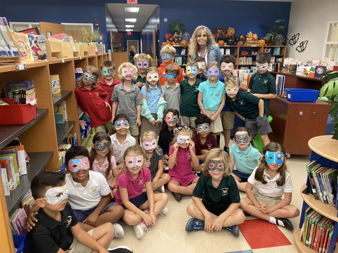 A large group of elementary students sit on the floor of a school library, each wearing a colorful, hand-drawn paper mask over their eyes. Their teacher stands behind them, smiling.