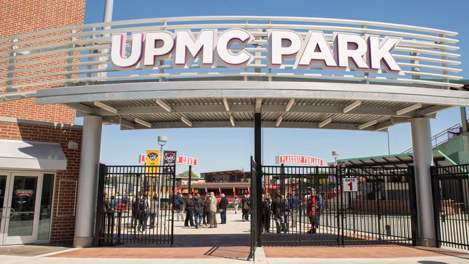 Entrance of UPMC Park with a large sign overhead. A group of people stands near the entrance, and various buildings and structures are visible inside.