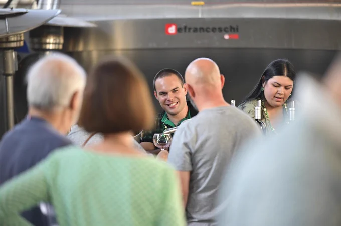 A gathering of people at a social event in front of a Defranceschi machine. Members of the group are engaged in conversation, with one person smiling broadly and another focused on a task. Bottles are visible on a counter in the background.