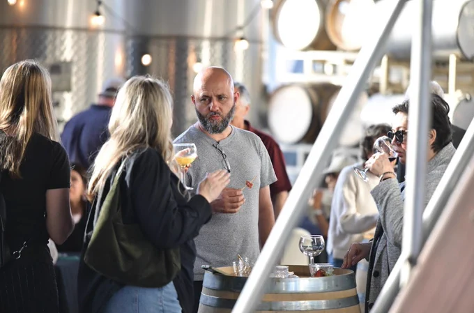 A group of people are gathered around a barrel table in a winery, engaging in conversation and drinking from wine glasses. The background shows large metal tanks and wooden barrels, typical of a brewery setting.