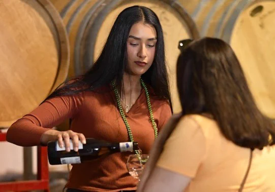 A person is pouring wine into a glass for another individual at a wine club pick-up event. Barrels are visible in the background.