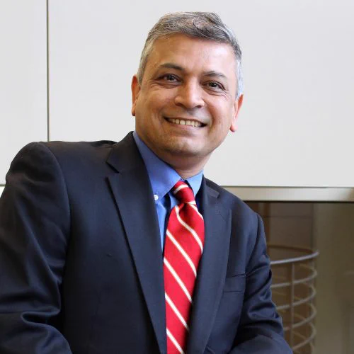 Man in a dark suit and red striped tie sitting indoors against a light-colored wall.