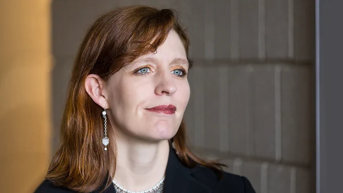Woman with shoulder-length brown hair wearing a black blazer and long dangling pearl earrings against a neutral background.