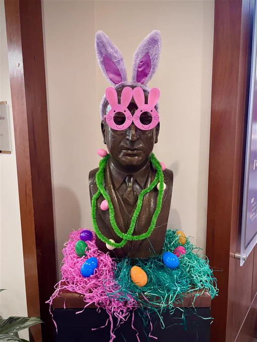 A bust of Arthur J. Morris wearing bunny ears, bunny glasses, and egg garland. The bust is surrounded by plastic grass and plastic eggs.