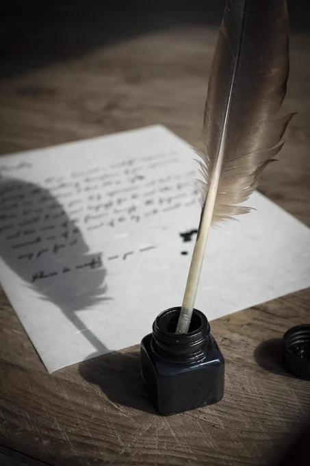 Eine Feder in einem Tintenfass vor einem beschrifteten Blatt Papier.