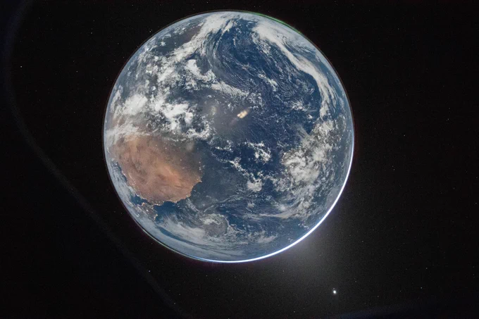 A full disc image of Earth, as seen from the Orion Crew Module. The planet is a pale blue, swirling with white clouds and glowing slightly lighter blue in place from reflected light. At lower left, a large brown landmass is Africa, with Spain and Portugal with twinkling lights where the planet curves. At top right, auroras glow in a thin green glow, just barely separated from the planet's surface. Earth is set against the black of space (pic: NASA/R.Wiseman)