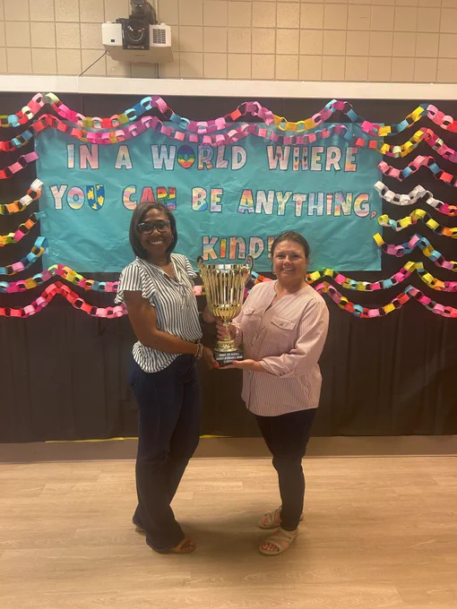 Two women holding a large trophy in front of a colorful bulletin board with a kindness-themed message.