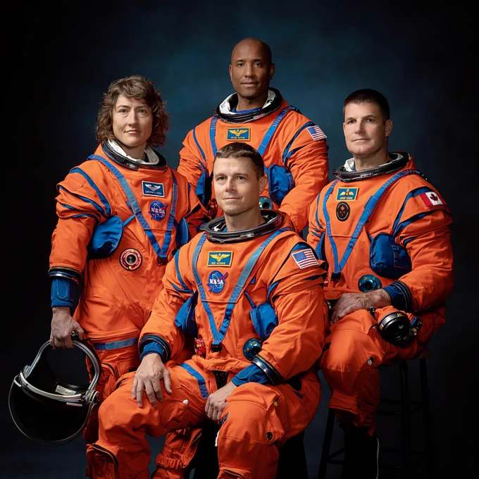 The four-person crew of the Artemis II mission posing in their orange Orion Crew Survival System pressure suits against a dark blue background. 

From left to right: NASA Mission Specialist Christina Koch, NASA Pilot Victor Glover (standing in the back), NASA Commander Reid Wiseman (seated in the front), and Canadian Space Agency Mission Specialist Jeremy Hansen.