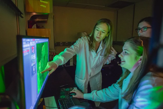 Sara Prescott points to a scientific image on a monitor screen in her lab while students Julia Van Goor and Emma Ibanez look on.