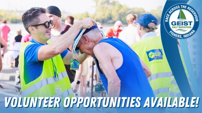 A person wearing a yellow vest places a medal on another individual at the Geist Half Marathon event. The background shows other participants and a body of water. Text on the image reads, "Volunteer Opportunities Available!" The event logo is displayed in the corner, saying "Geist Half Marathon, Race Across the Reservoir, Fishers, Indiana."
