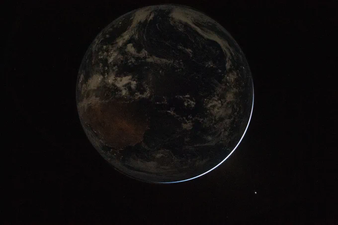 Earth as seen from the Orion capsule. It is divided sharply in half, with a line running diagonally from the 11 o'clock to the 4 o'clock. The lower left half is a bright pale blue. Its surface is covered with swirly, puffy white clouds, with texture indicating different thicknesses. The upper right half is dark, with no illumination from the Sun. Earth is set against the black of space. Credit: NASA/Reid Wiseman
