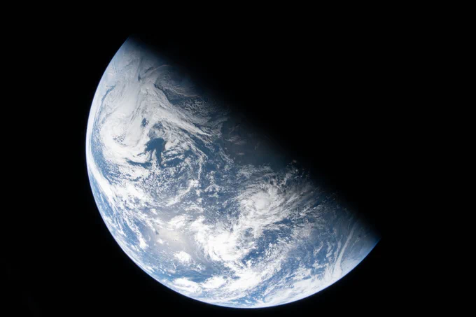 Earth as seen from the Orion capsule. It is divided sharply in half, with a line running diagonally from the 11 o'clock to the 4 o'clock. The lower left half is a bright pale blue. Its surface is covered with swirly, puffy white clouds, with texture indicating different thicknesses. The upper right half is dark, with no illumination from the Sun. Earth is set against the black of space. Credit: NASA/Reid Weisman