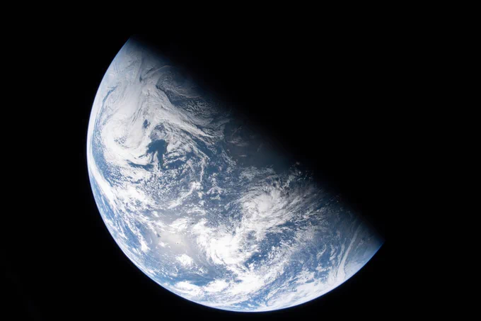 Earth as seen from the Orion capsule. It is divided sharply in half, with a line running diagonally from the 11 o'clock to the 4 o'clock. The lower left half is a bright pale blue. Its surface is covered with swirly, puffy white clouds, with texture indicating different thicknesses. The upper right half is dark, with no illumination from the Sun. Earth is set against the black of space. Credit: NASA/Reid Wiseman