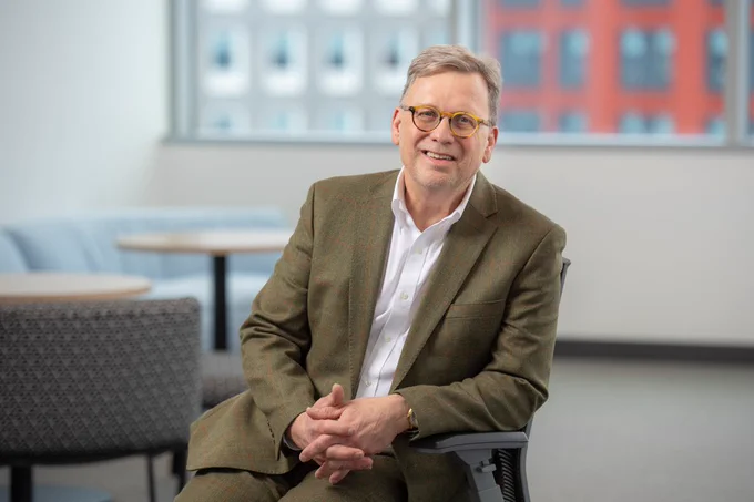 Dean Lindstrom with glasses and a green tweed blazer, sitting in an office chair with his hands clasped. He is smiling warmly in a bright, modern office setting with blurred furniture and windows in the background.