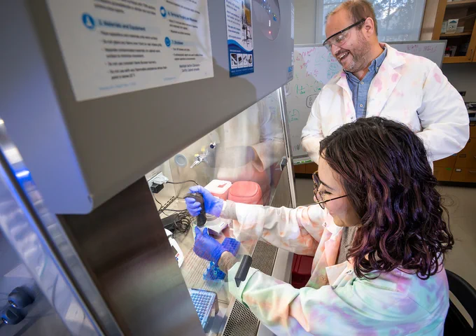 A professor looks over a student's shoulder as she handles samples in a protected lab shelf.