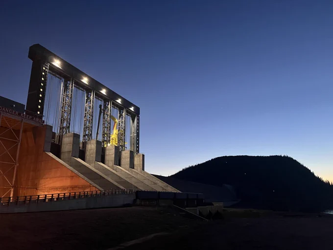 The image shows a dam illuminated by artificial lights against a dusk sky. The structure features large hydraulic gates and is positioned next to a dark silhouette of a hillside in the background.