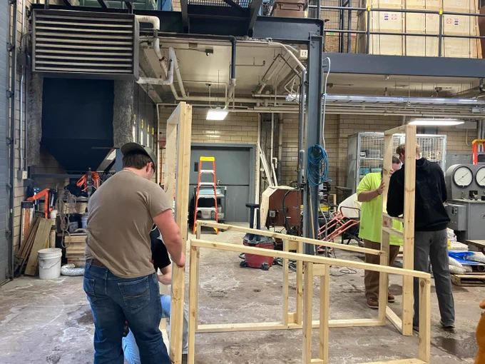 Students assemble a wooden frame structure in a lab, positioning vertical and horizontal beams while working together to align components. Tools, ladders, and equipment are visible in the background.