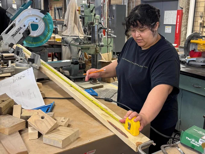 A student measures a long wooden board with a tape measure at a workbench near a miter saw. Wood blocks and tools are scattered around, highlighting precise fabrication work.
