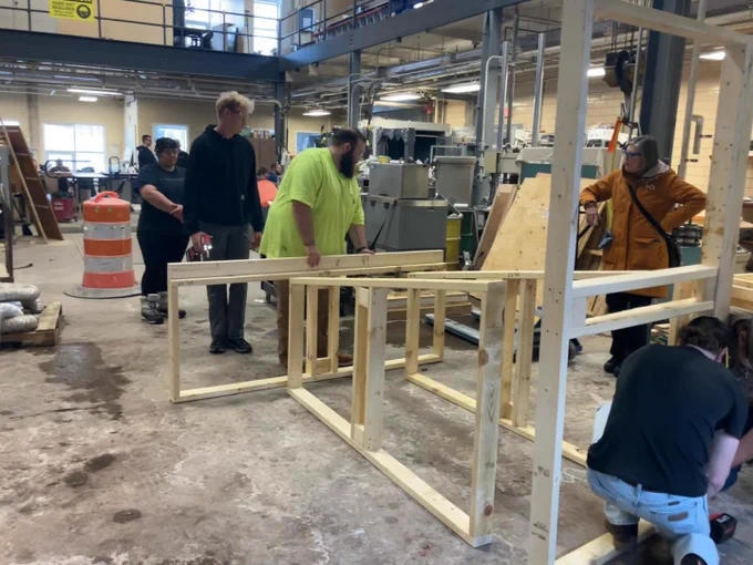 A group of students works together to construct a multi-section timber frame structure on a workshop floor while an instructor observes. The partially assembled structure shows progress toward a full-scale build.