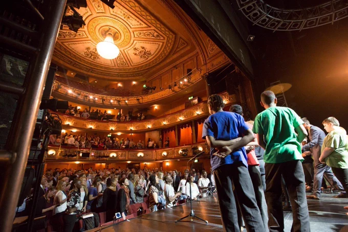 Children on a theatre stage, receiving a standing ovation from an audience.