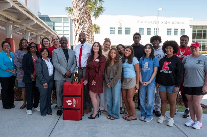A diverse group of people, including adults and students, are gathered in front of Edgewater High School. They are standing on a sidewalk with palm trees and the school building visible in the background. One person holds a red bag with a logo.