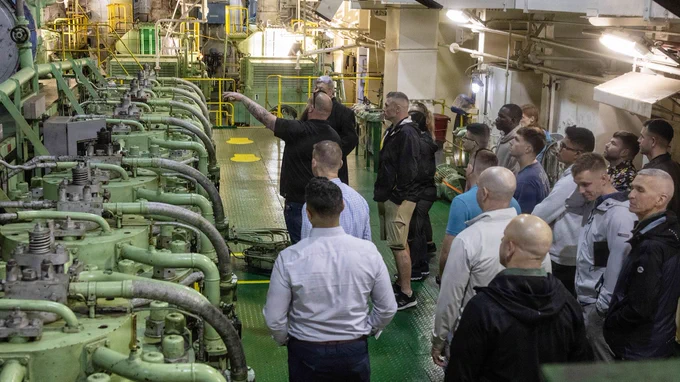 Marines and civilians tour an engine room aboard a Military Sealift Command vessel, observing machinery as a crew member explains operations.