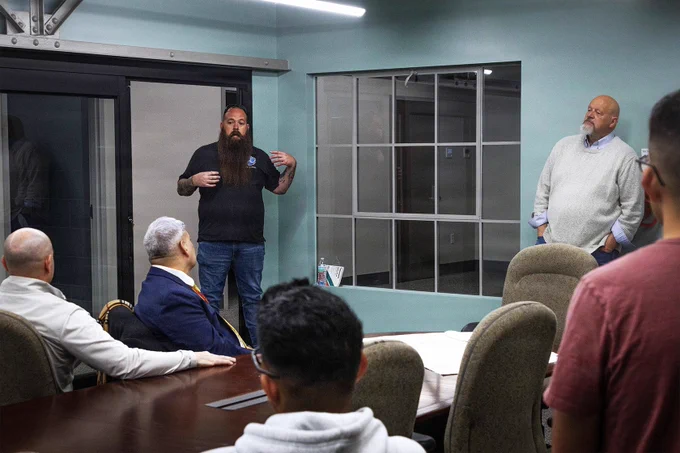 Marines and civilians receive a briefing in a shipboard conference room, discussing operations and capabilities during a vessel visit.