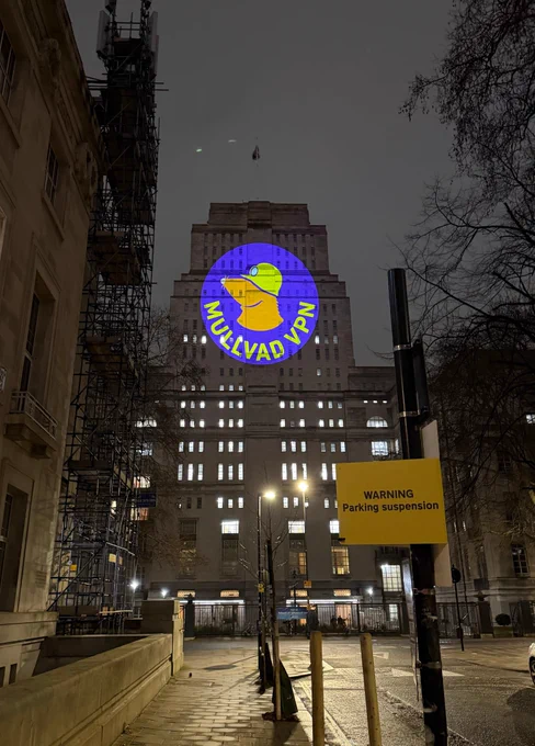 A photograph of Senate House in London during the night time. 

Projected onto the front of it is the Mullvad VPN logo in a circle with a purple background, the words Mullvad VPN are written in yellow across it.