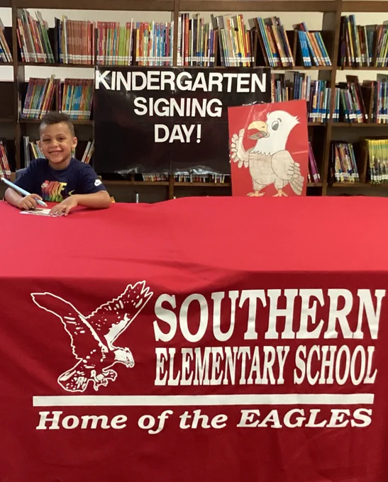 A young child signing a document at a red tablecloth-covered table with Southern Elementary School and eagle logo, celebrating Kindergarten Signing Day in a library.