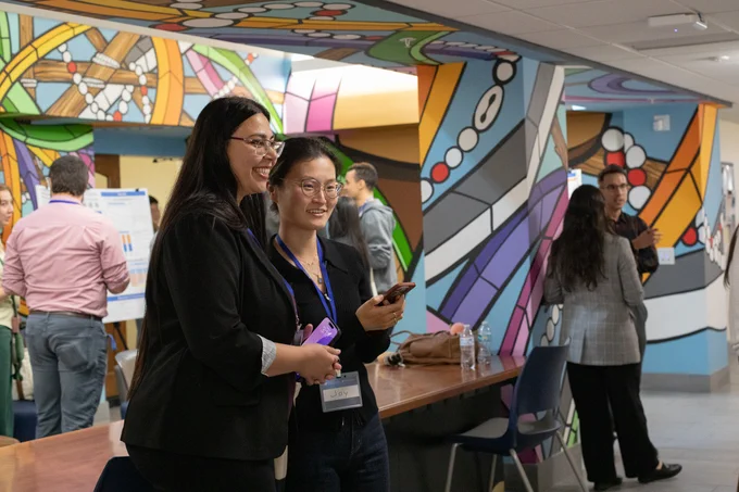 Two attendees of the National AI Literacy Day @ UF smile while looking at something that is off camera, presumably an academic research poster.