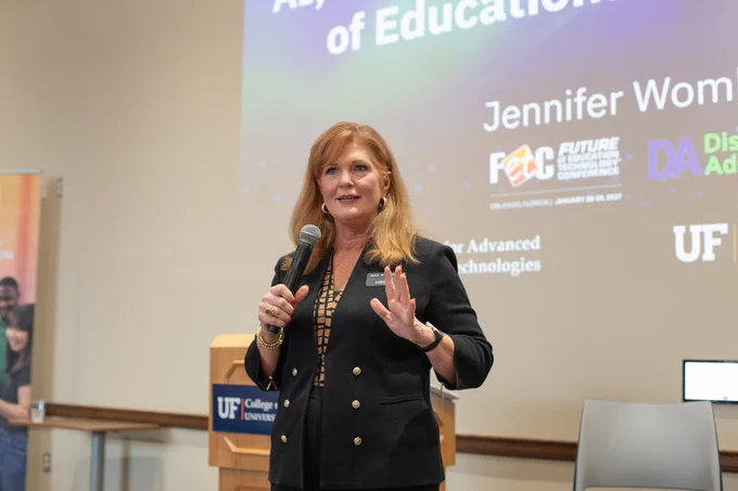 Jennifer Womble from FETC gives the keynote at the National AI Literacy Day @ UF conference. She is wearing a navy blazer and speaking enthusiastically in the Norman Conference Center. 