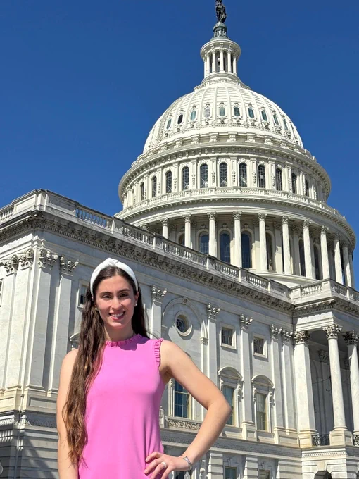 A person in a pink top stands smiling in front of the US Capitol building on a clear day.
