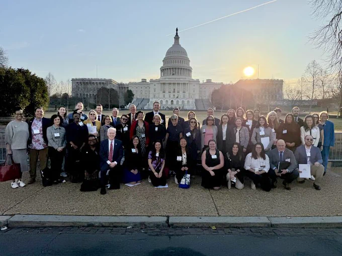 A large group of people posing in front of the United States Capitol building during sunset. Some are standing while others are sitting in the front row. The sky is clear with a setting sun. A barricade is visible behind them.