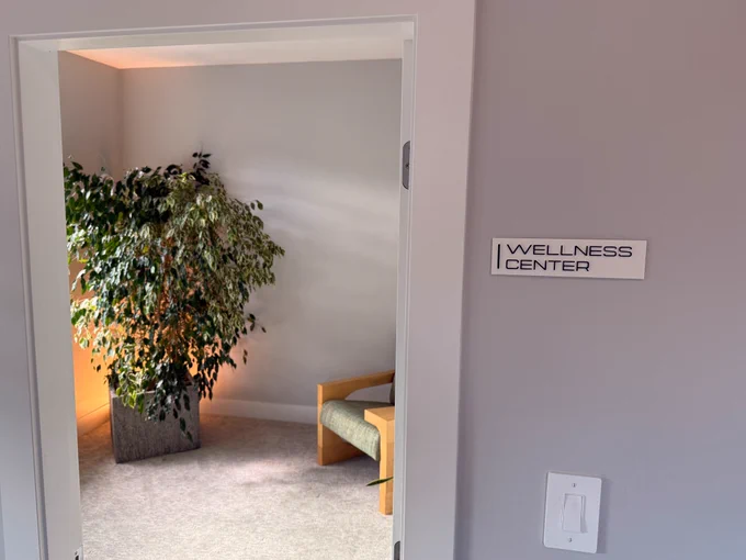 The photo depicts a doorway into a room with a fig tree and a single chair. To the right of the doorway there is a plaque that has "Wellness Center" written on it.