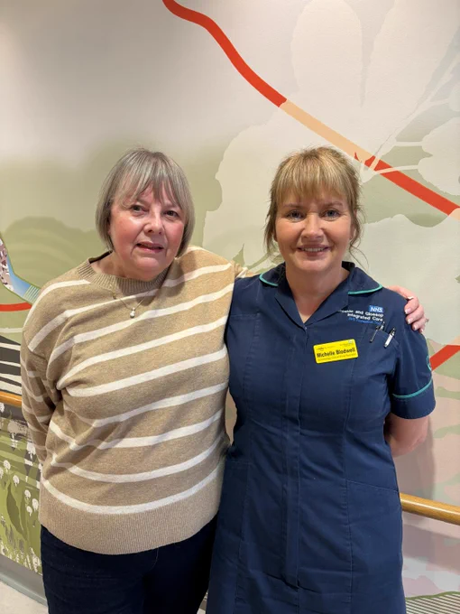 Catherine a woman with bob length grey hair wearing a stripy jump stands in a hospital setting with nurse Michelle wearing a medical uniform