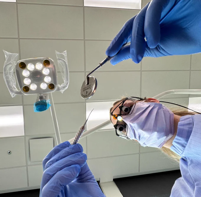 A dental student from RCC wearing protective gear and a headlamp is holding dental tools, preparing for a procedure. The perspective is from the patient's viewpoint, looking up at a ceiling light in a dental office.
